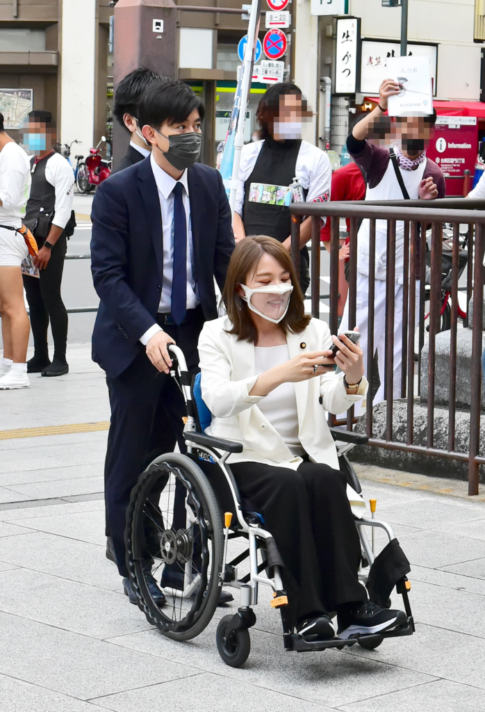 骨折して車椅子の今井絵理子議員のサポートをする橋本健さん